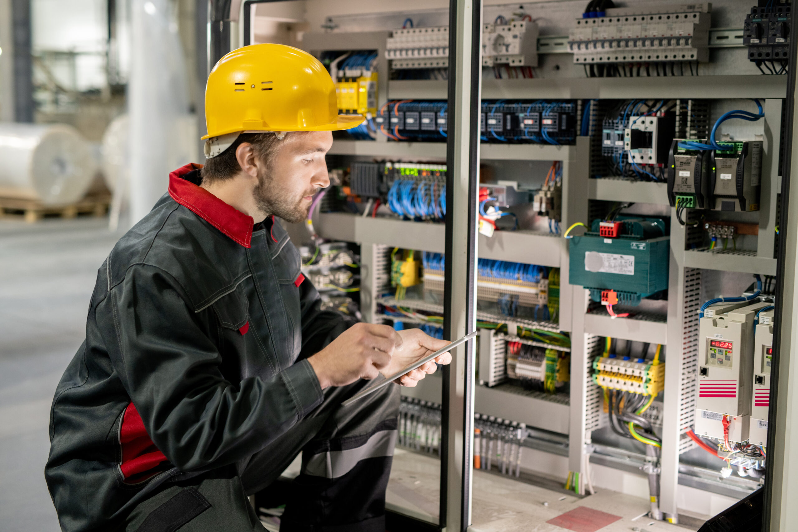 Contemporary young engineer in workwear and hardhat scrolling through data in tablet while squatting by one of industrial machines